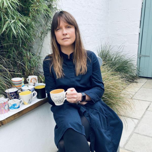 Young white woman with brown hair and a blue dress sits outside with a cup of tea 