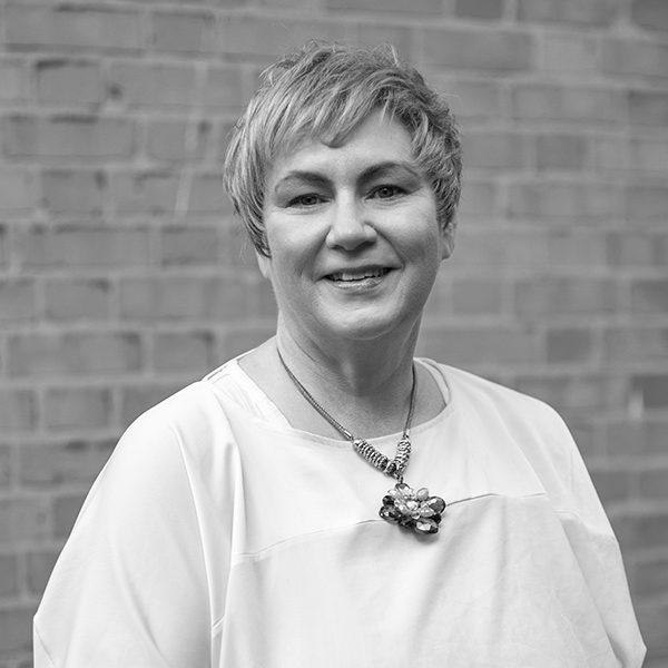 White woman with short fair hair in a white tshirt stands in front of a brick wall