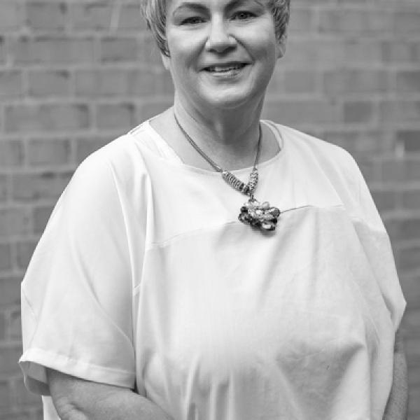 White woman with short fair hair in a white tshirt stands in front of a brick wall